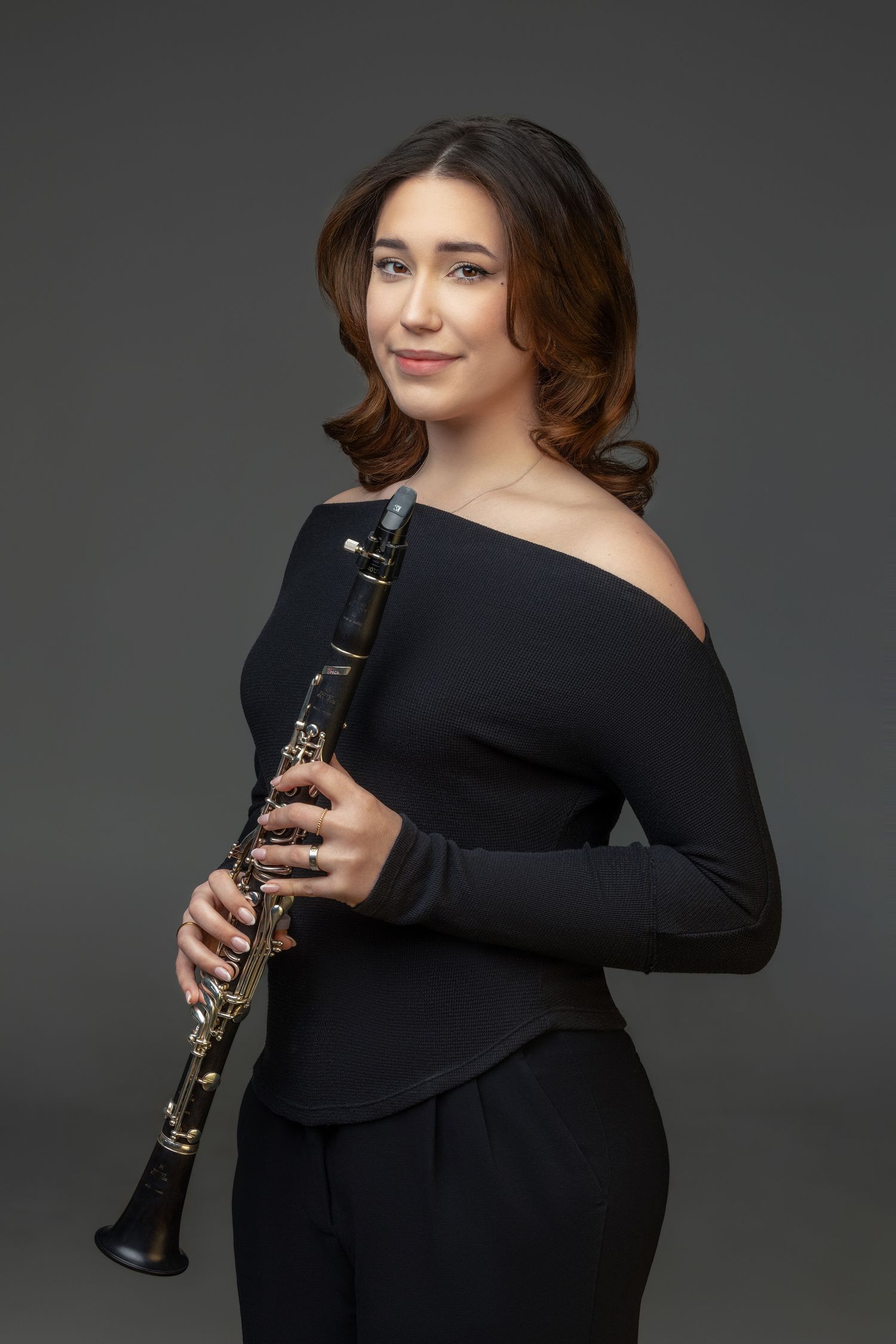 Professional musician poses in black off-shoulder dress holding a clarinet against dark studio background.