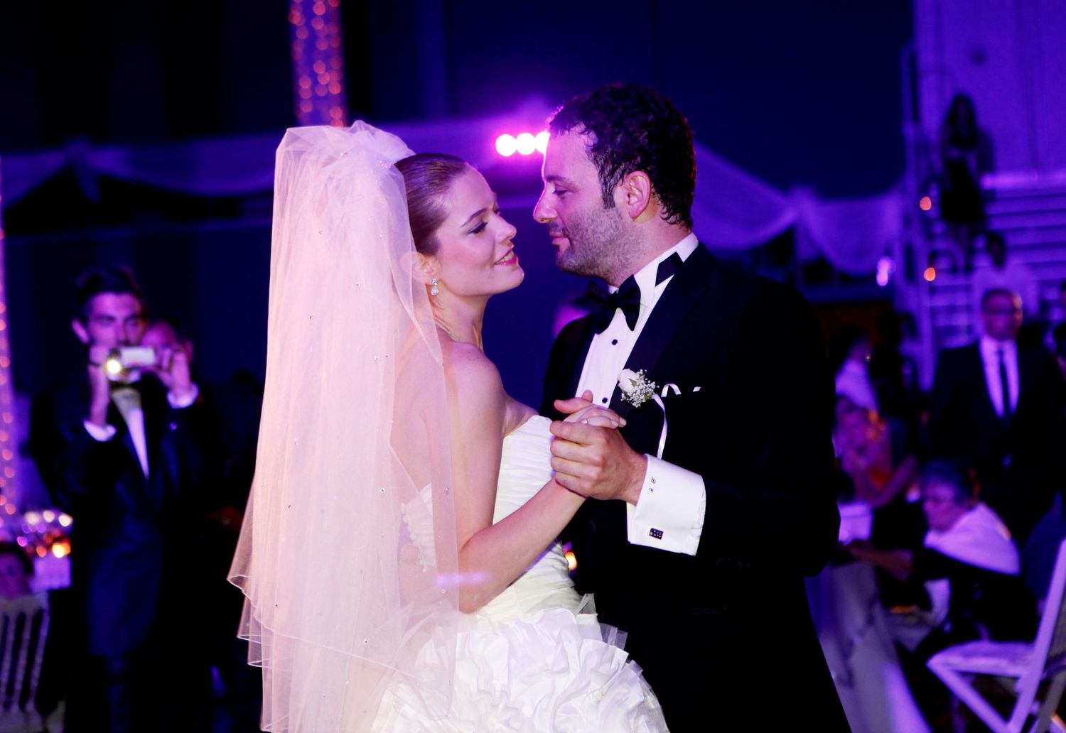 Newlywed couple sharing their first dance at a wedding reception with purple mood lighting and romantic atmosphere.