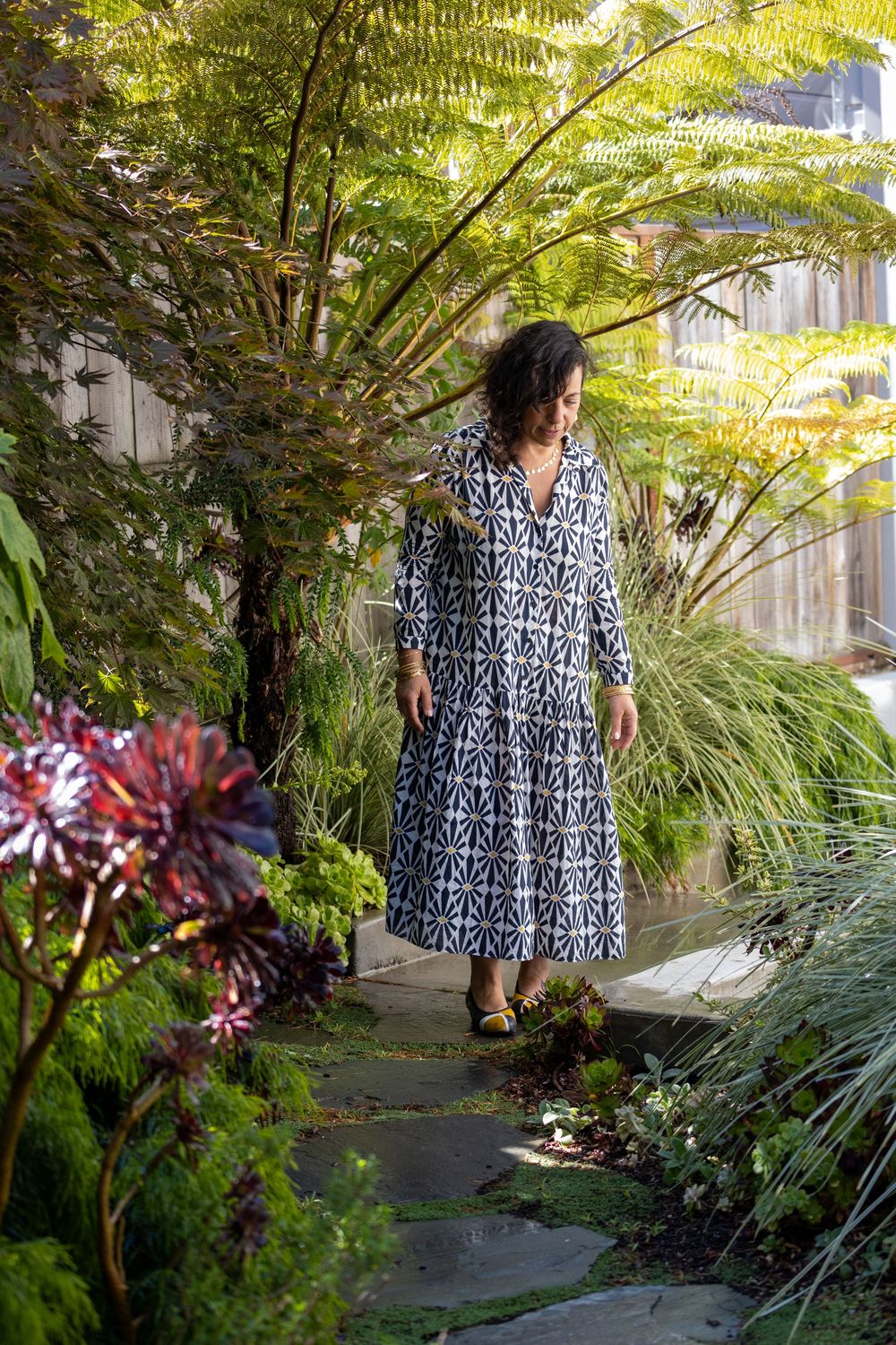 A woman walks along a garden path surrounded by lush ferns and tropical plants while wearing a patterned maxi dress.