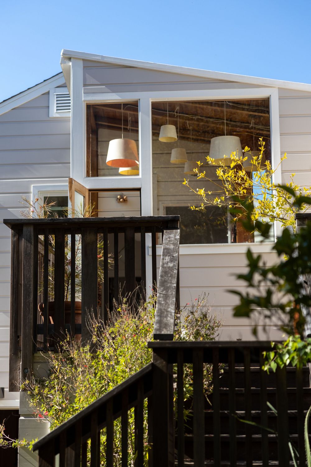 Black outdoor deck railing against white siding with hanging pendant lights and yellow flowering plants.