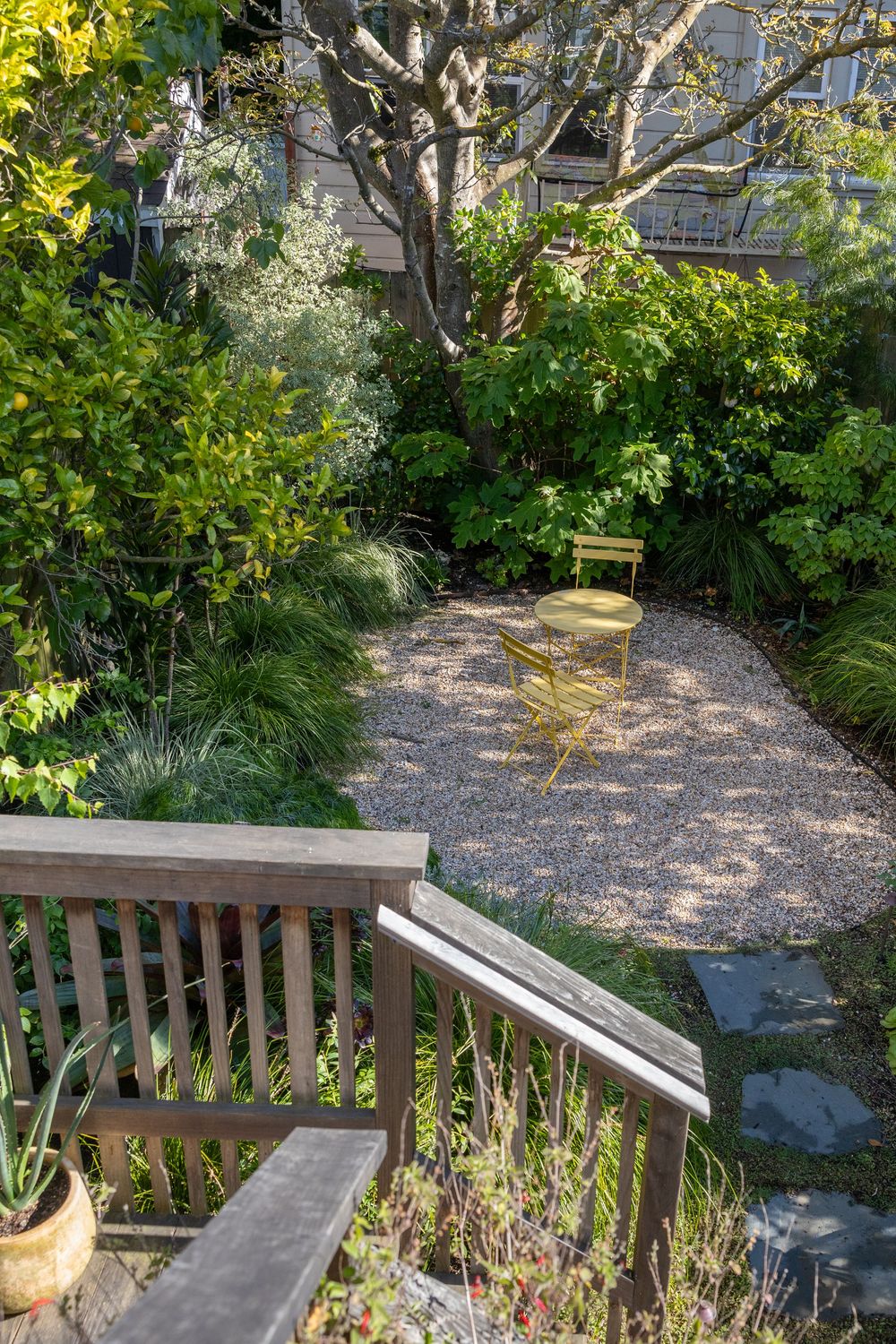 A peaceful garden path with gravel leads to a yellow metal chair surrounded by lush greenery and trees.