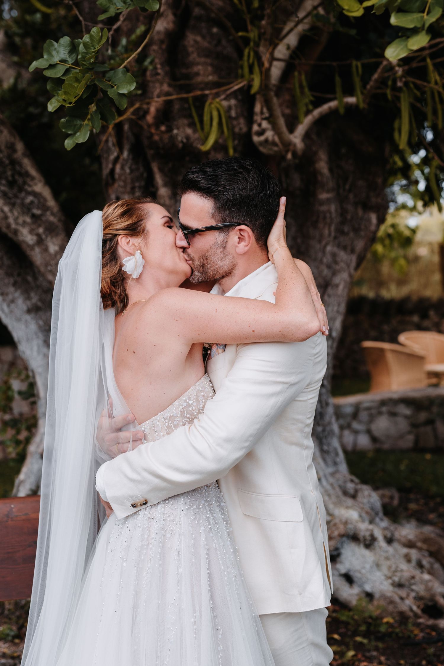 Romantisches Hochzeitspaar umarmt und küsst sich im Freien in einem Garten mit üppigem Grün im Hintergrund auf der Finca Xiclati, Mallorca.