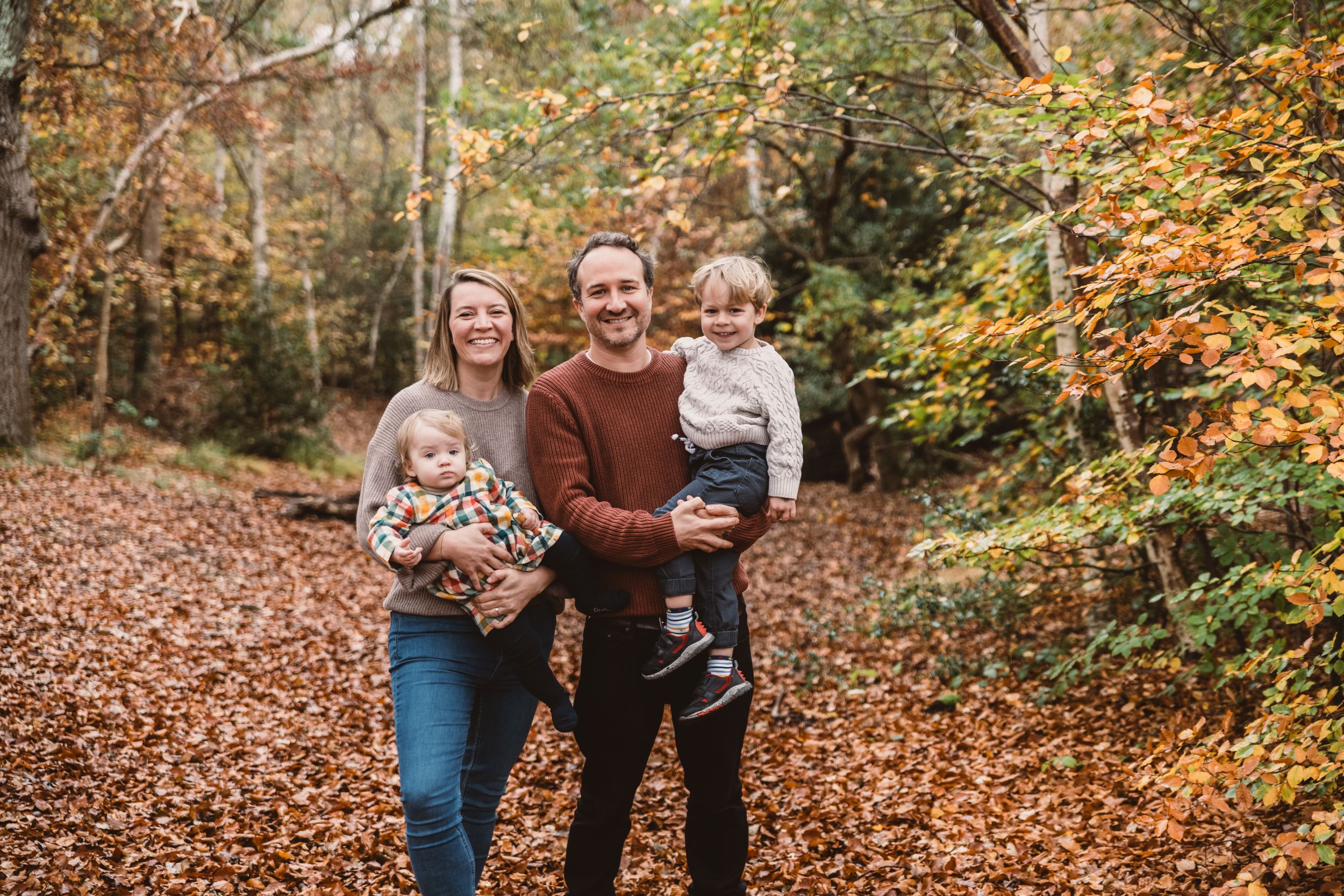 Family walking and playing together on a path covered in fallen autumn leaves in a forest setting.