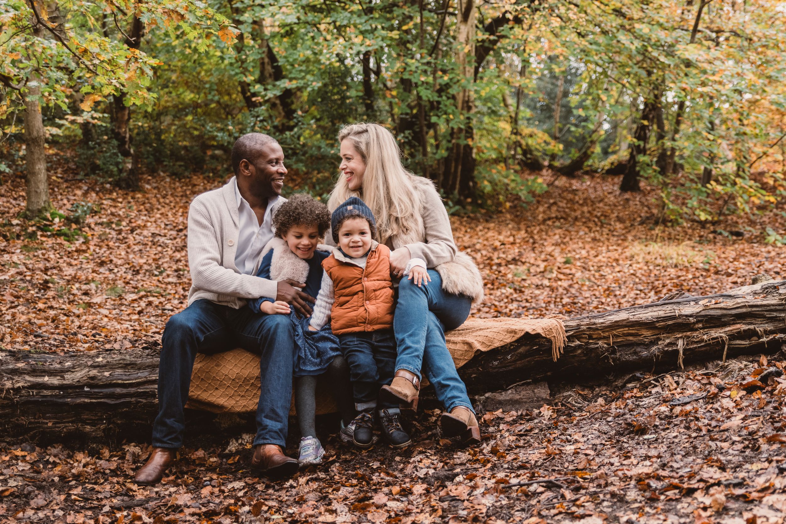 A family sits together on a fallen log surrounded by autumn leaves in a forest setting.