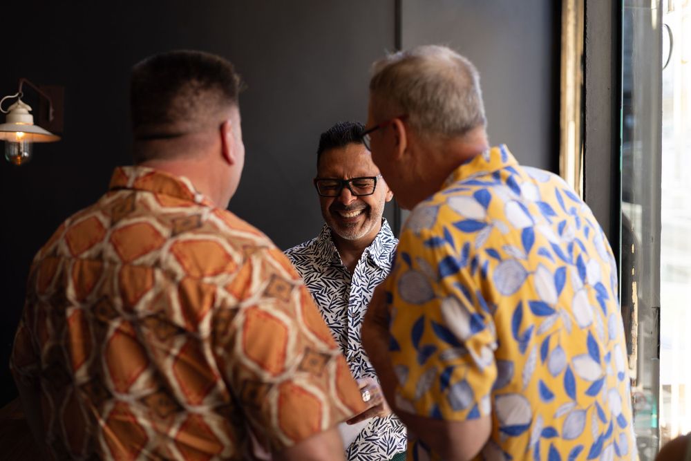 Two people in patterned shirts sit at a bar counter, enjoying conversation and drinks in an indoor setting.