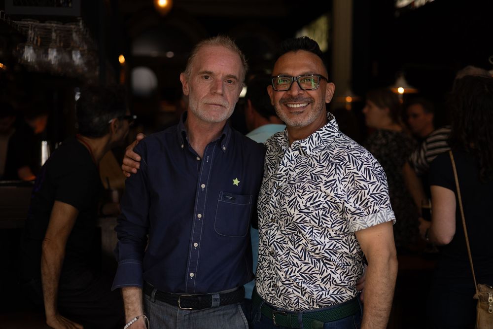 Two people smiling together at a nighttime event, one wearing a blue button-down and one in a patterned short sleeve shirt.