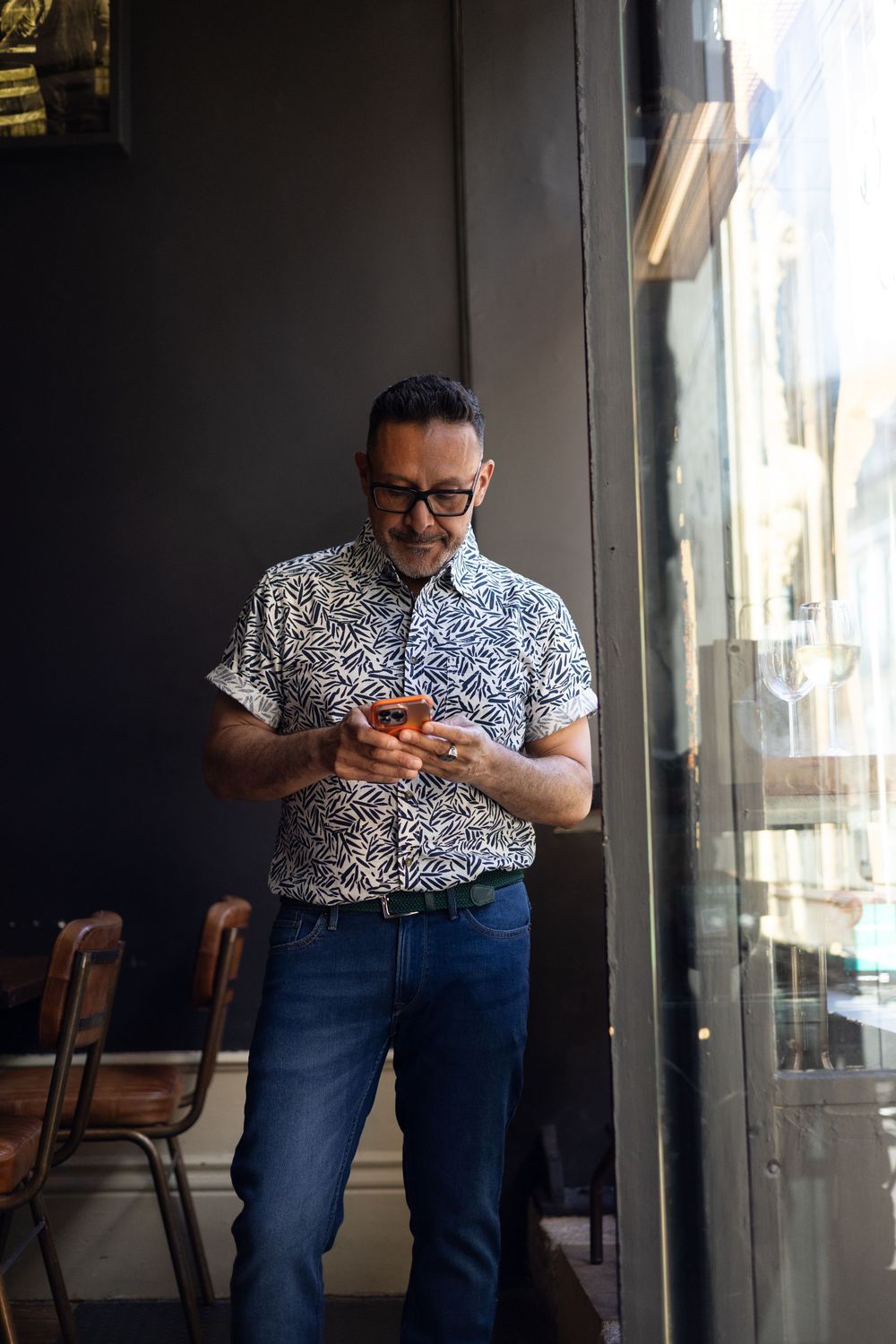 Someone in a patterned shirt and jeans is shown looking at their phone in dark lighting conditions.