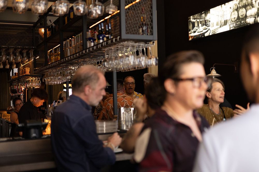 Patrons socialize at a dimly lit bar with glassware hanging overhead in a cozy cocktail establishment.