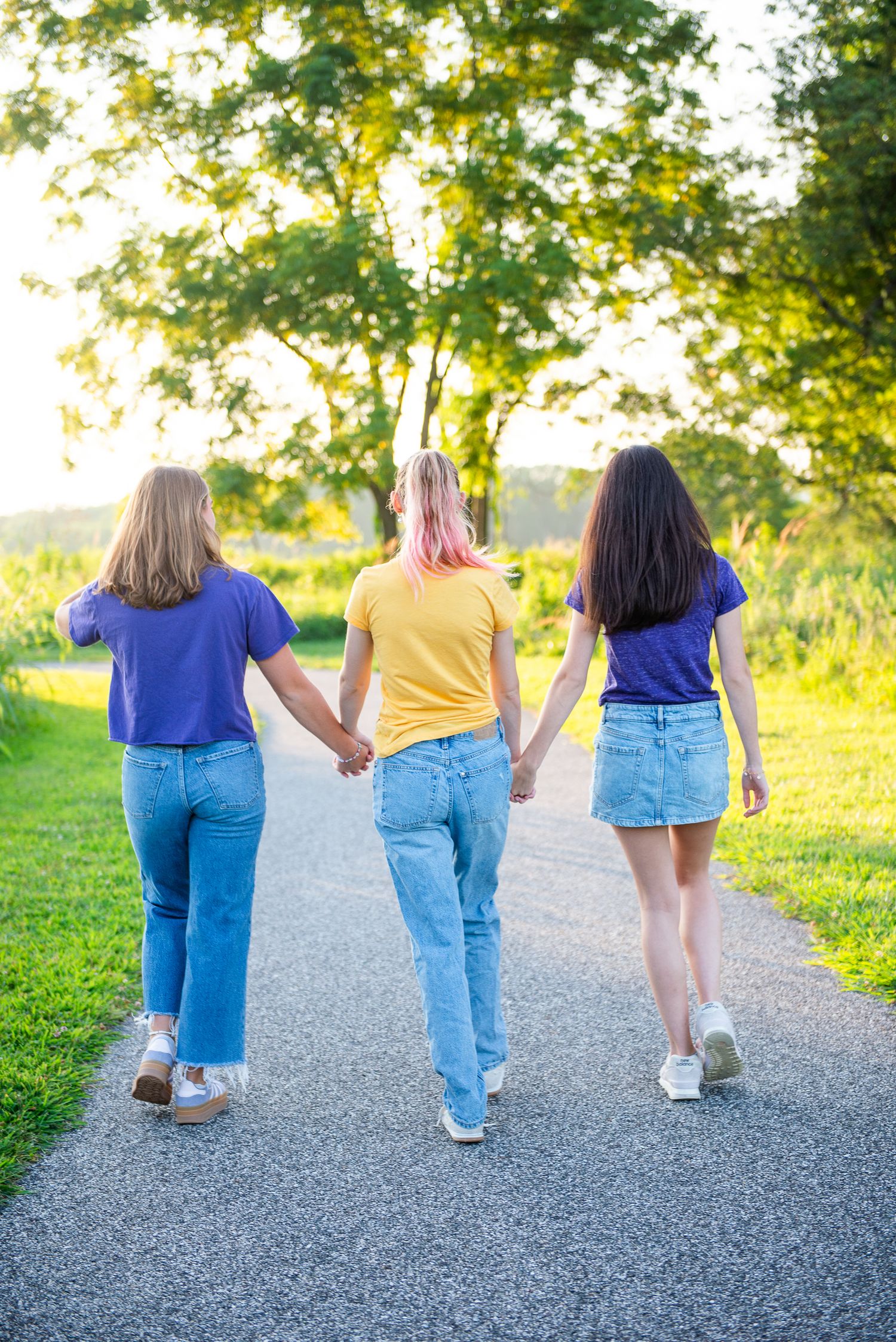 Three friends walking together on a paved path surrounded by lush greenery on a sunny summer day.