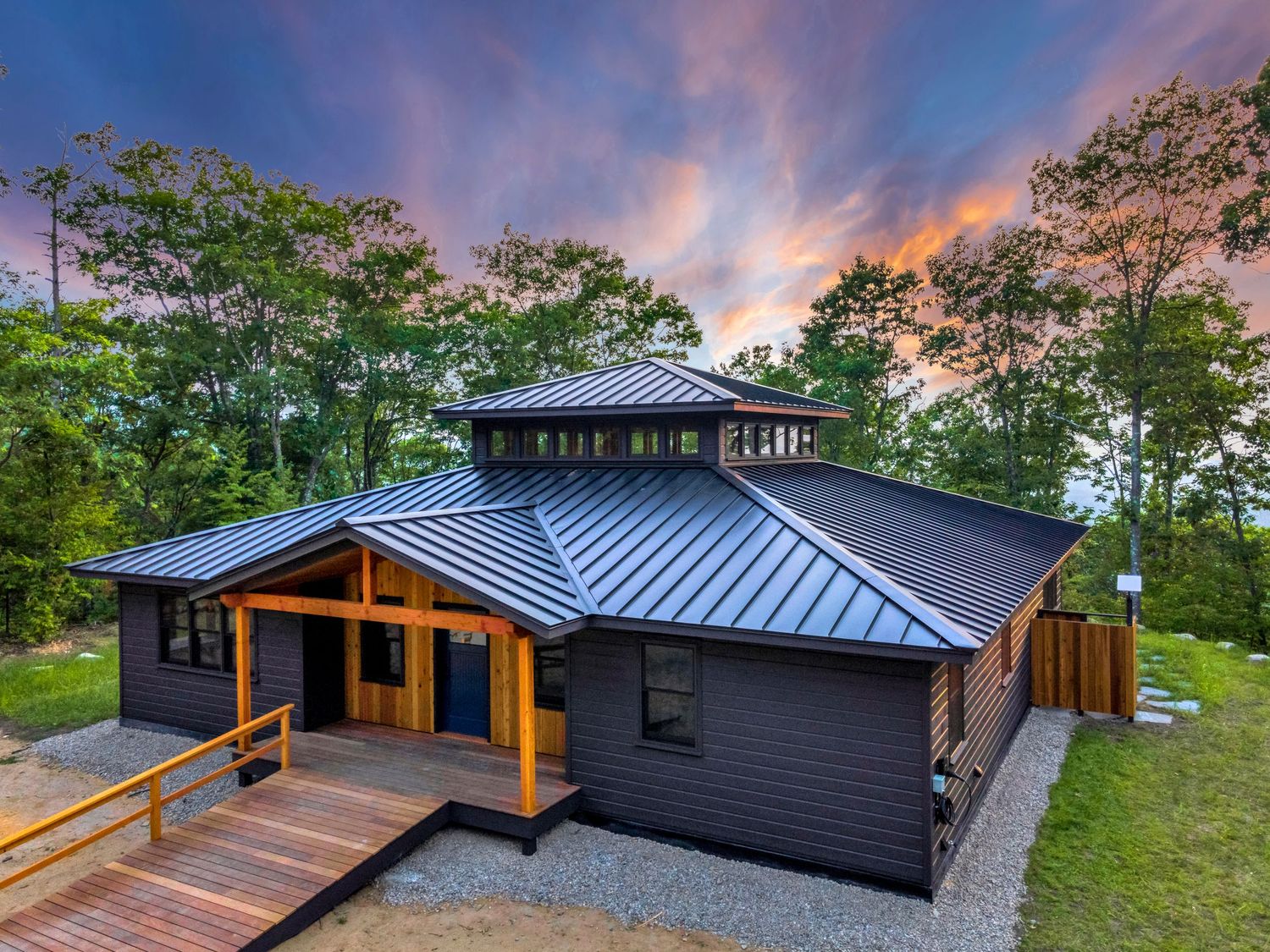 Modern dark gray house with metal roof and wooden deck features dramatic sunset sky and surrounding green trees.