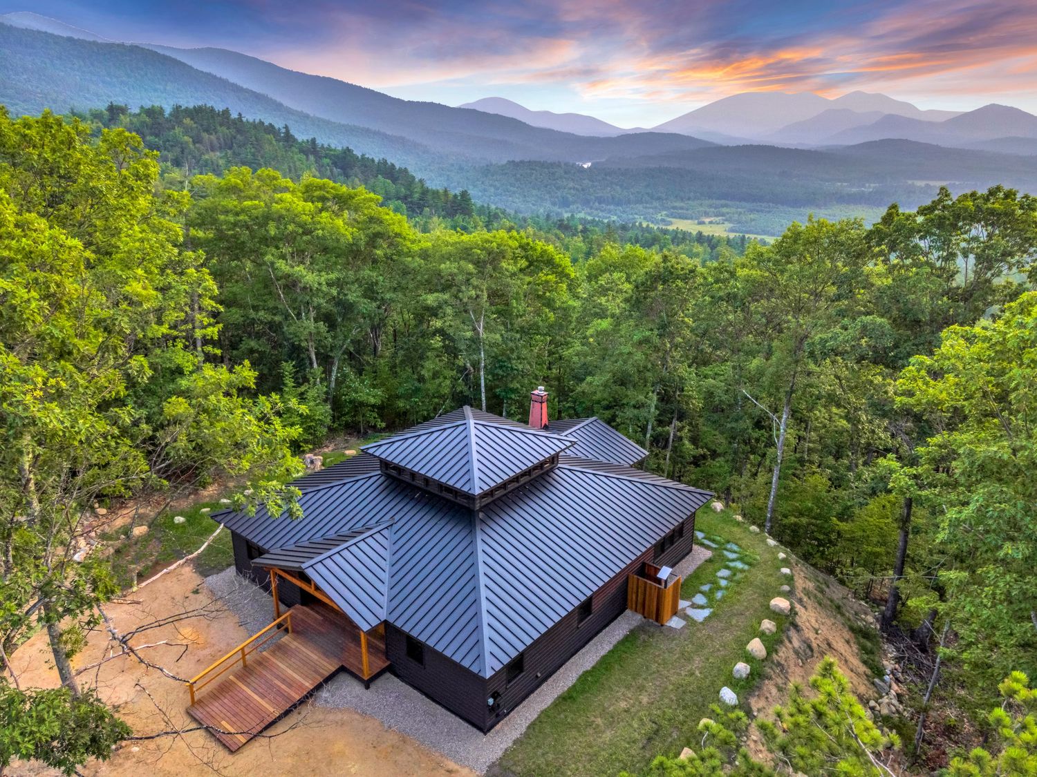 Modern mountain cabin with blue metal roof nestled in lush forest overlooking scenic Blue Ridge Mountains at sunset.