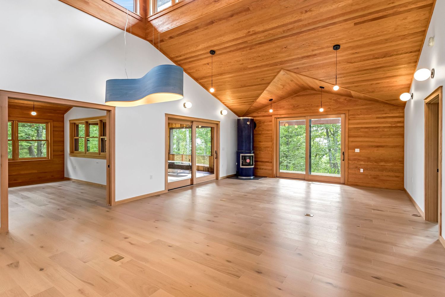 Modern open concept living room with vaulted wood ceiling, hardwood floors, and large windows overlooking nature.