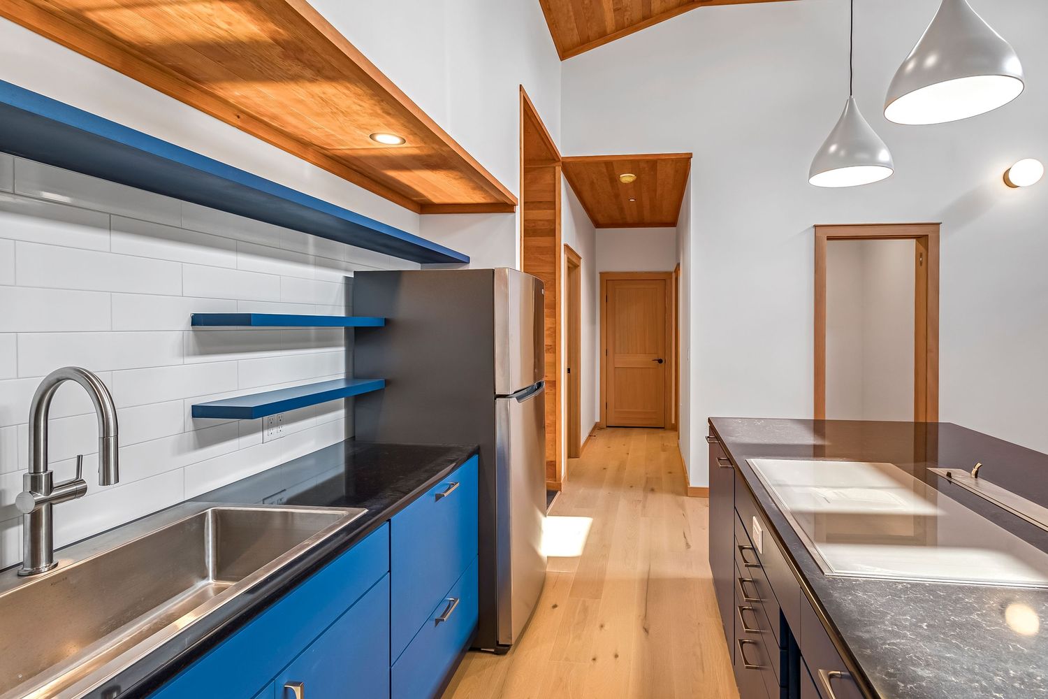 Modern kitchen with blue cabinets, wooden beams, and pendant lights features sleek stainless steel appliances and white counters.