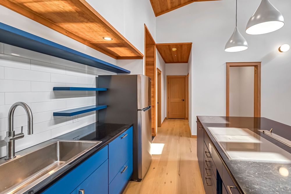 Modern kitchen with blue cabinets, wooden beams, and pendant lights features sleek stainless steel appliances and white counters.