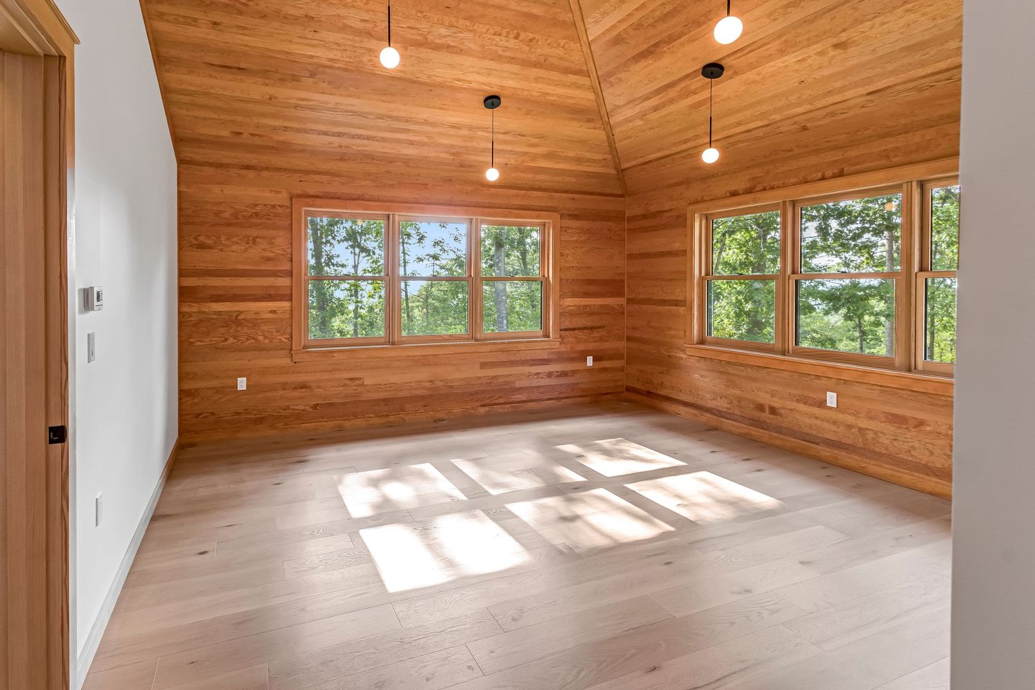 Bright empty room with wood paneled walls, large windows, light hardwood floors and exposed wood ceiling with pendant lights.
