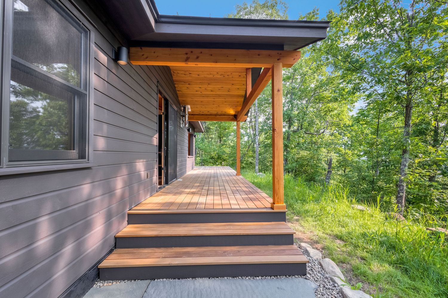 Modern home exterior with covered wood deck patio and stairs leading to lush green surroundings.