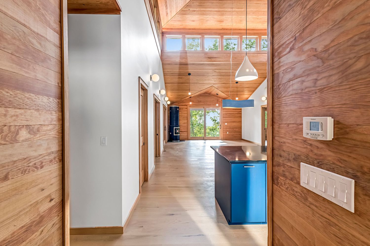 Modern hallway with warm wood paneled walls, light oak flooring, and blue cabinetry leads to an open living space.