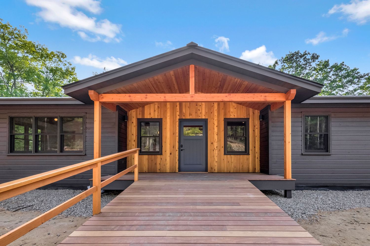 Modern cabin exterior with wooden porch, ramp entrance, and gray siding under blue sky.