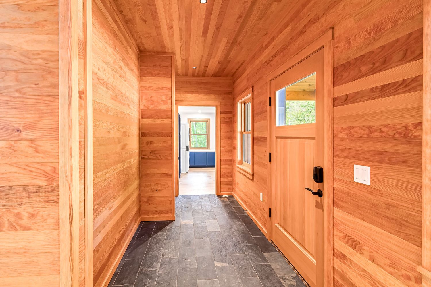 Sunlit corridor with natural wood wall paneling, slate floor tiles and a wooden door features contemporary finishes.