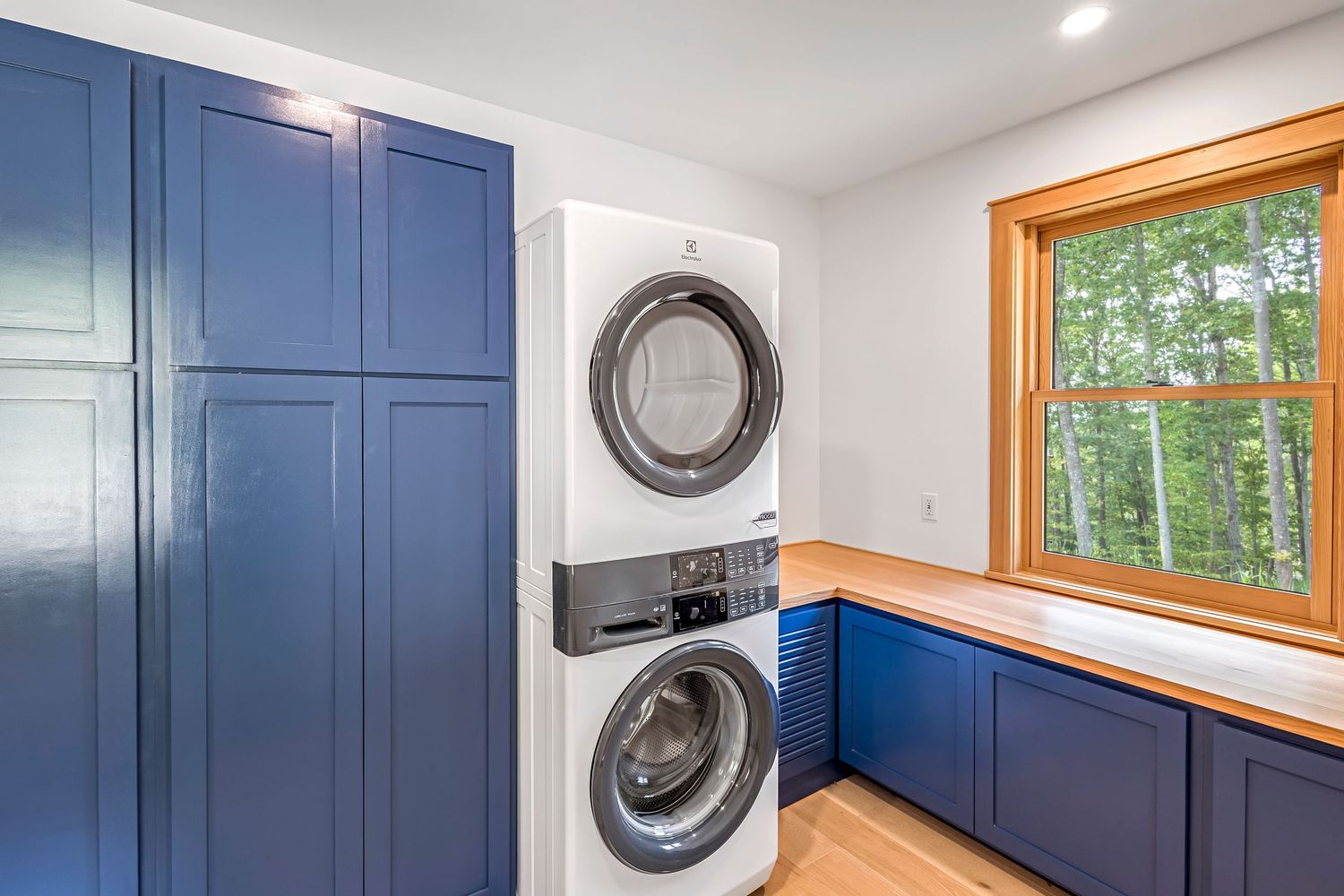 Contemporary laundry room with stacked washer-dryer, navy blue cabinets, and natural light from window.
