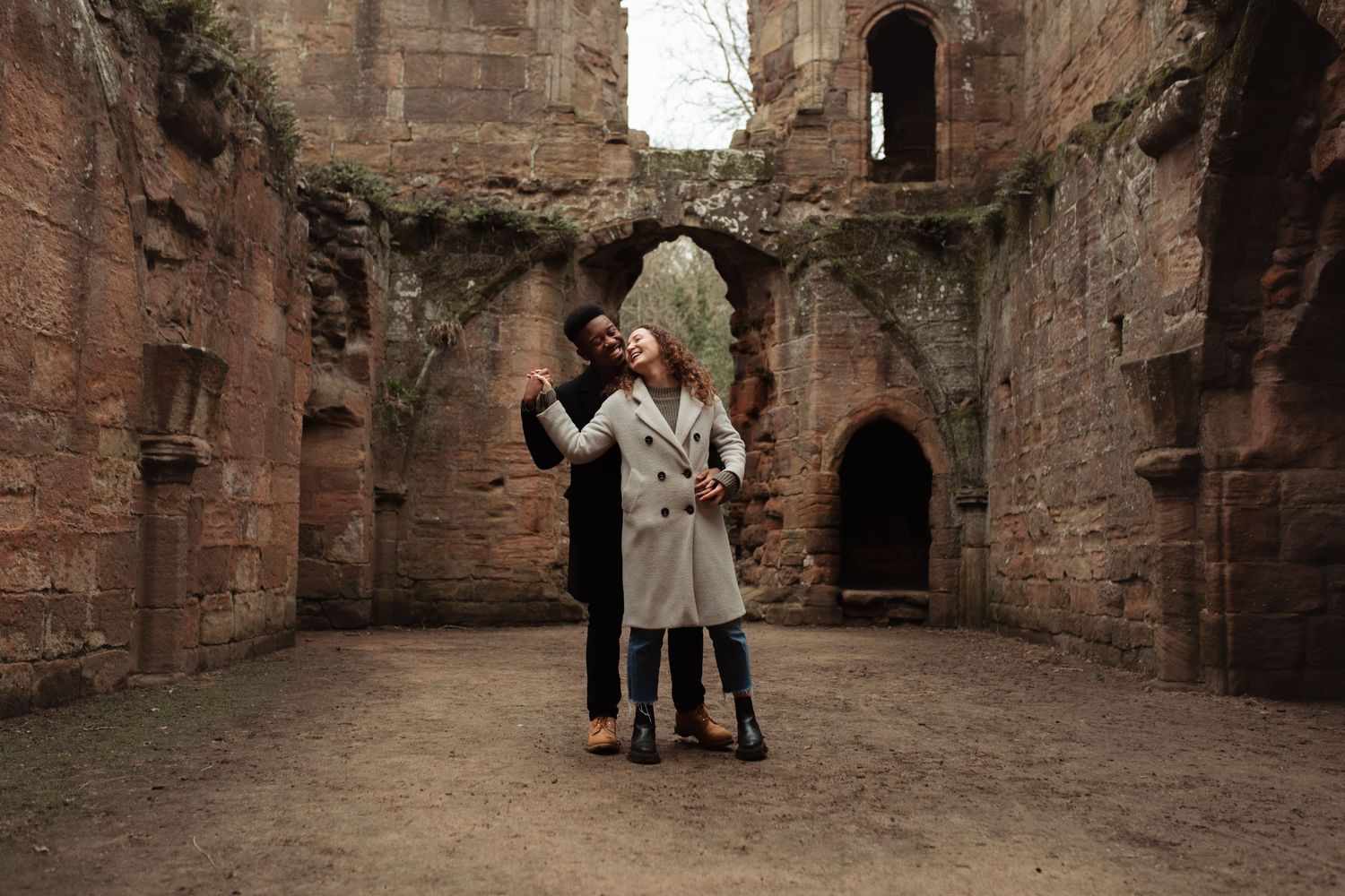 Couple sharing a romantic embrace inside ancient stone castle ruins with weathered red brick walls and arched windows. Harrogate Photo.