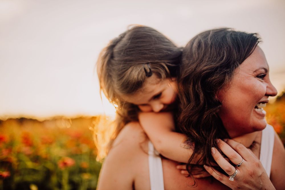A joyful mother laughs as her young daughter hugs her from behind in a sunny flower field at golden hour.