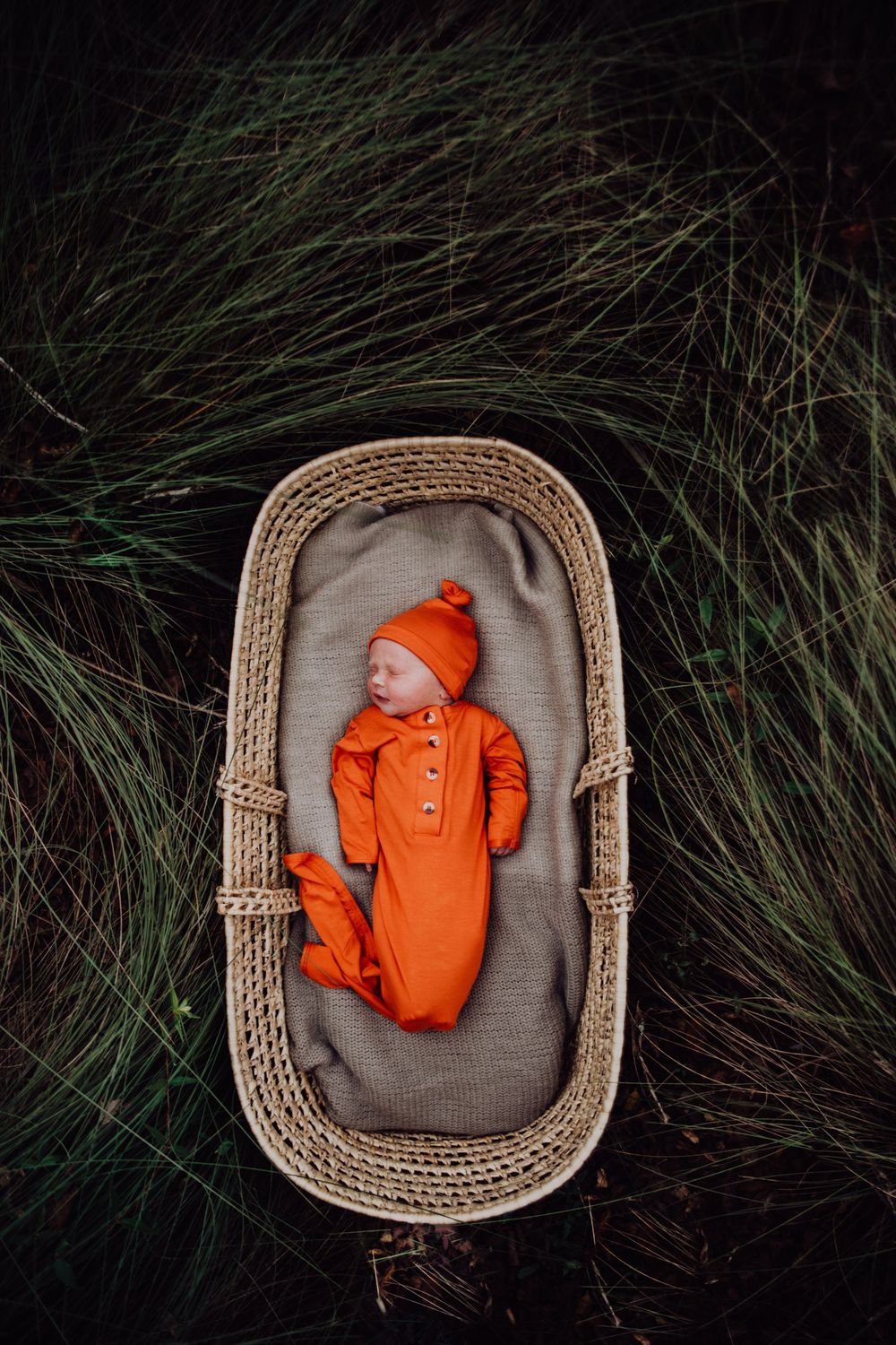 Small woven basket with sleeping baby wearing orange outfit and hat laying on gray blanket surrounded by green grass.