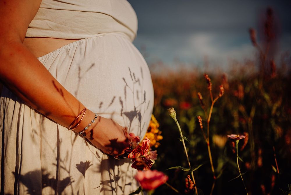 Pregnant woman in white dress holding wildflowers in a sunlit field, showcasing a beautiful maternity outdoor photo.