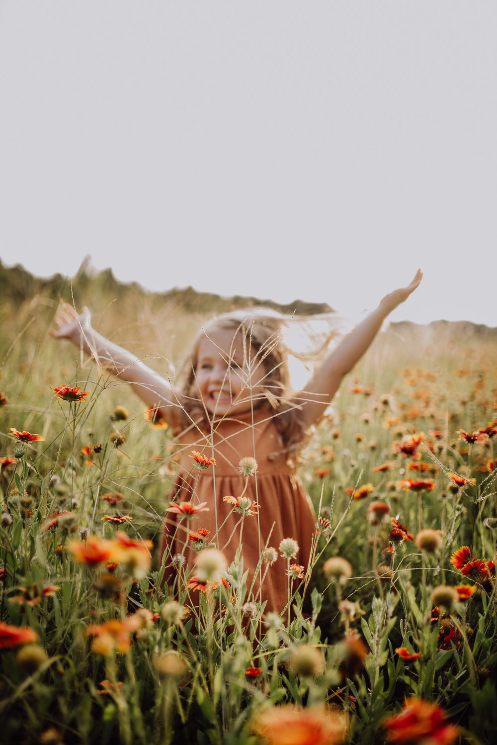 A person wearing a brown dress stands joyfully with raised arms among vibrant orange wildflowers in a summer meadow.