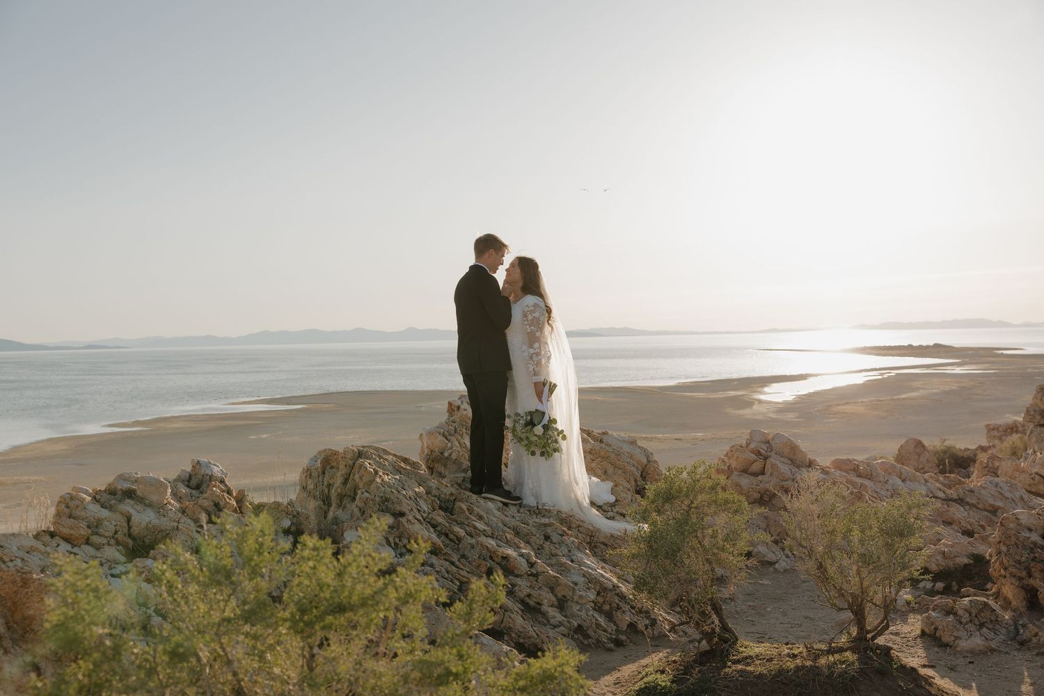Bride and groom standing on rocky overlook with vast lake and sandy shoreline in background.