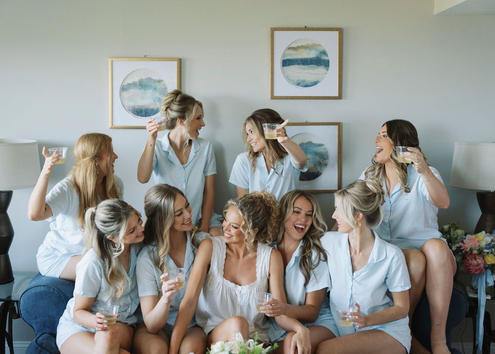 Bridesmaids in matching blue robes laughing and drinking champagne together before the wedding.
