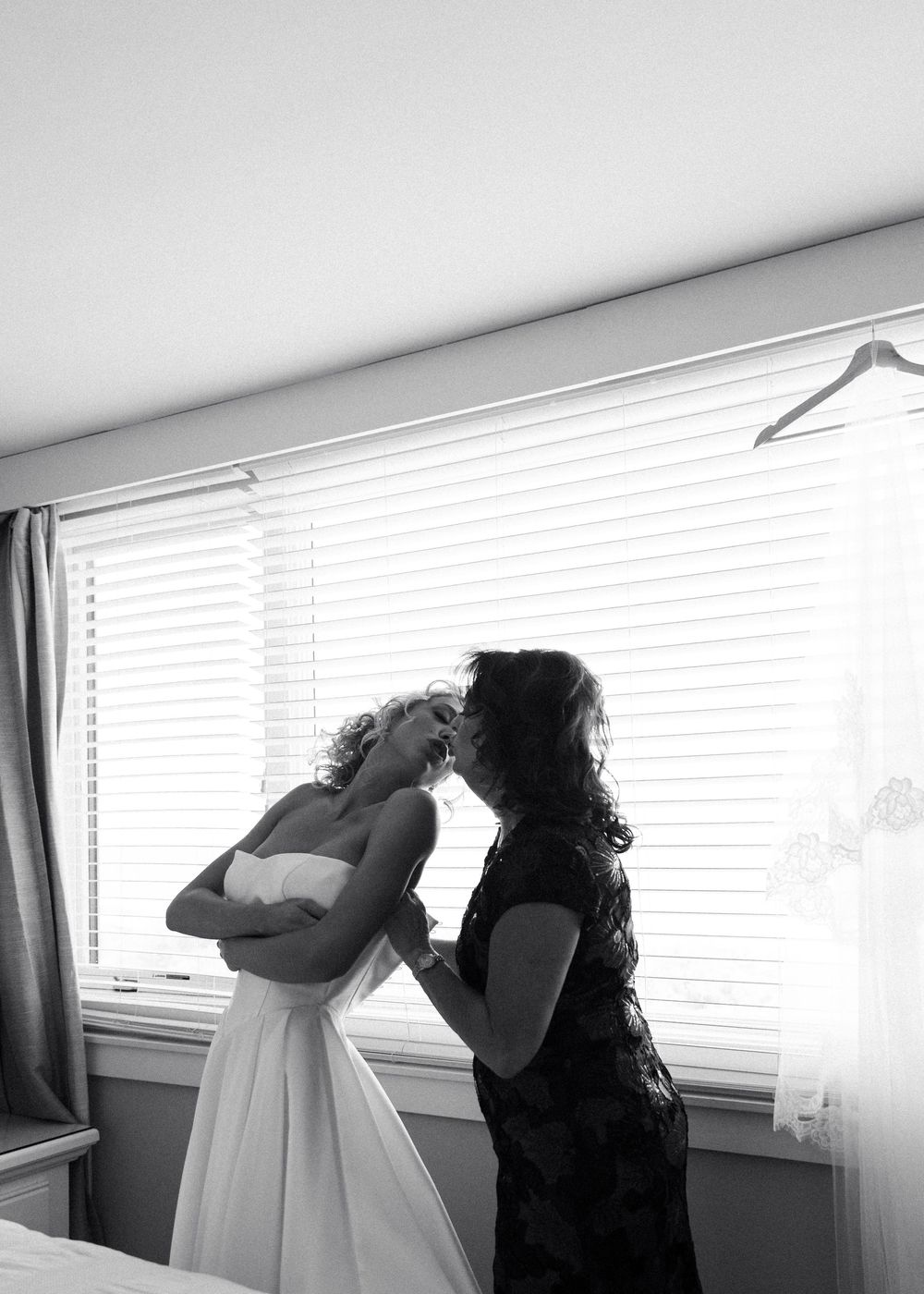 Black and white photo of mother helping bride into wedding dress by sunlit window.