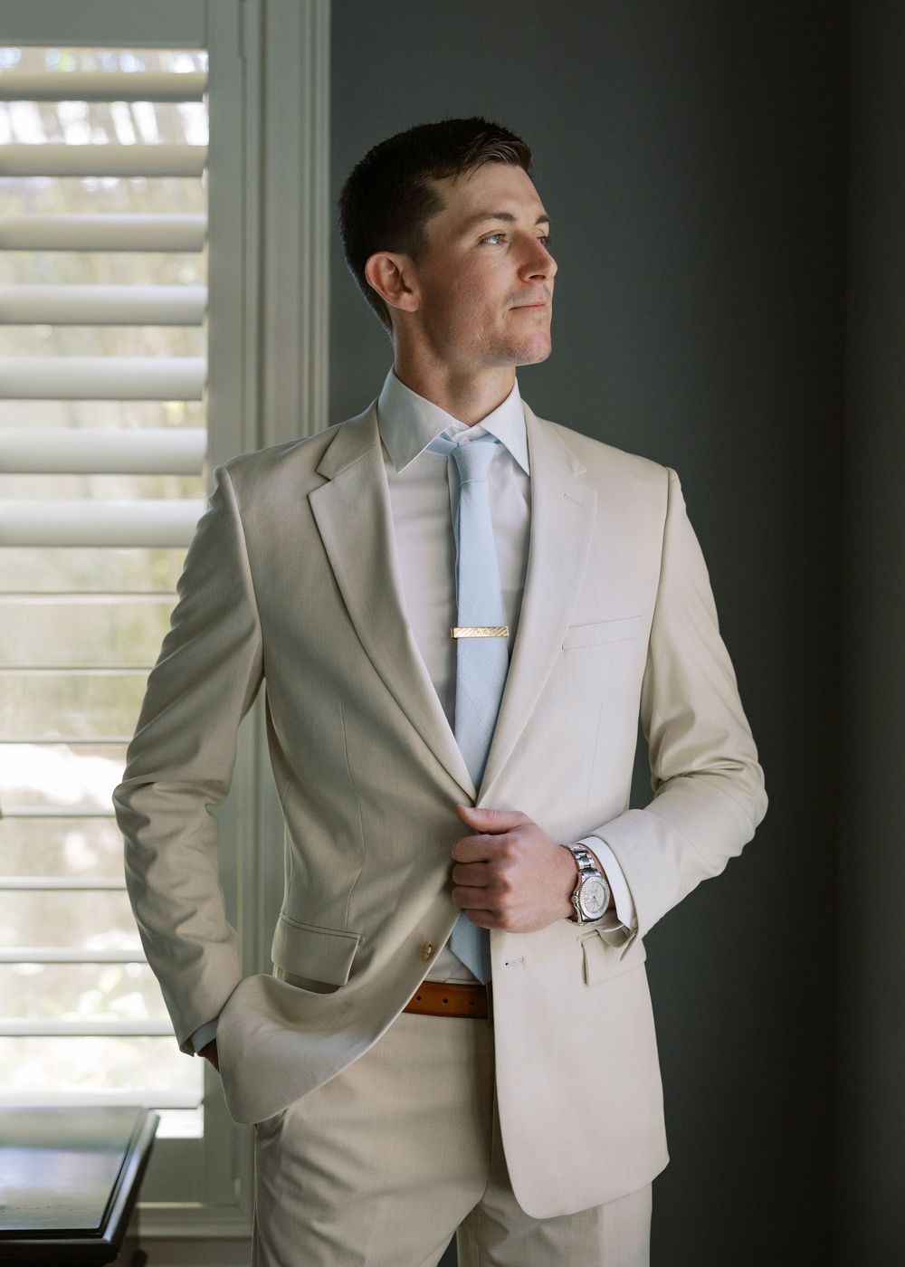 Dapper groom in a tan suit and light blue tie gazes out a window before his wedding.