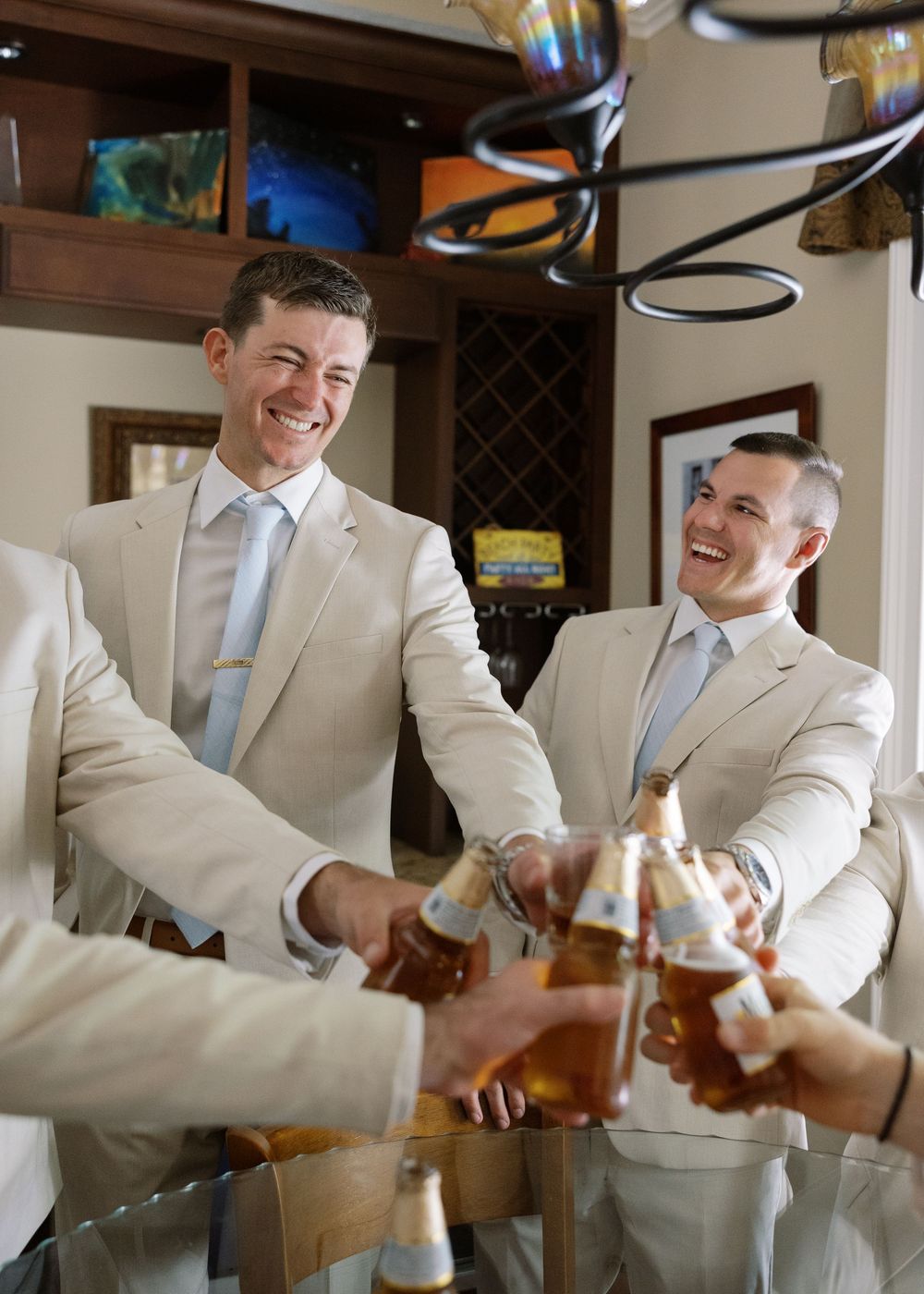 Groomsmen in tan suits clink beer bottles together in a toast at a wedding celebration.
