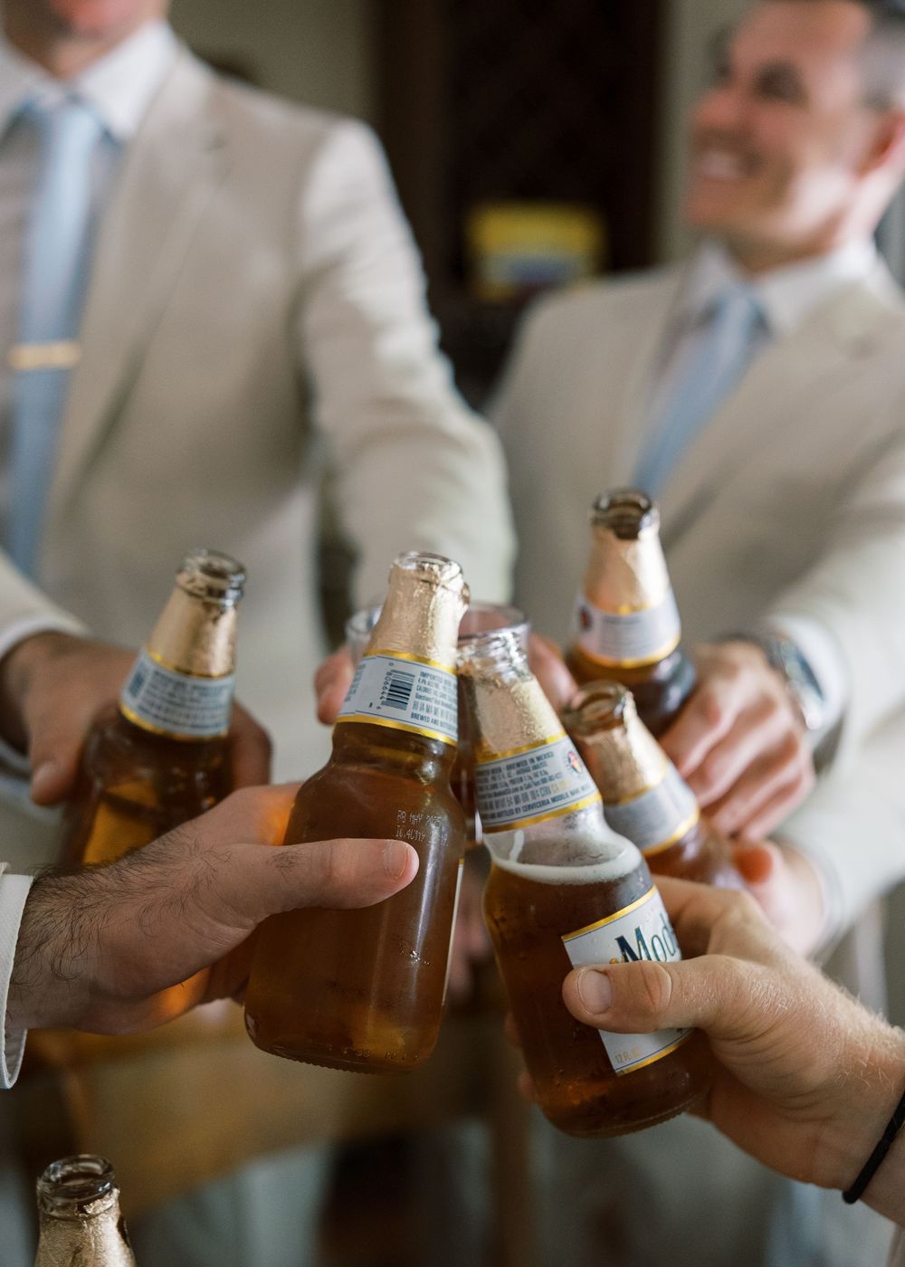 Groomsmen in beige suits clinking beer bottles together in celebratory pre-wedding toast.