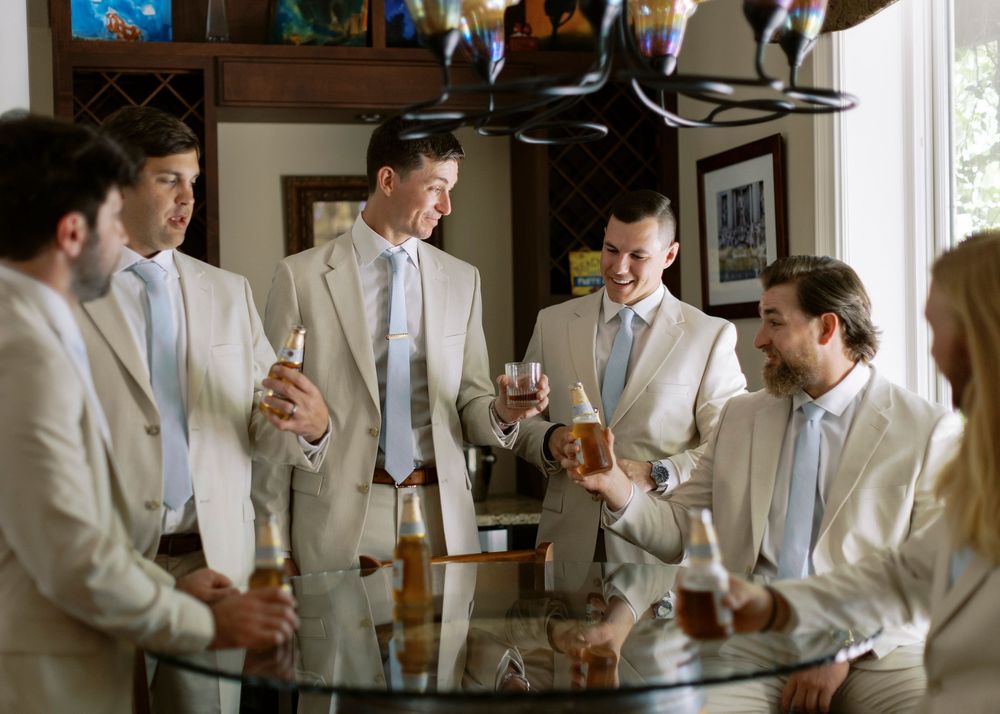 Groomsmen in tan suits gather around a glass table sharing drinks before the wedding ceremony.