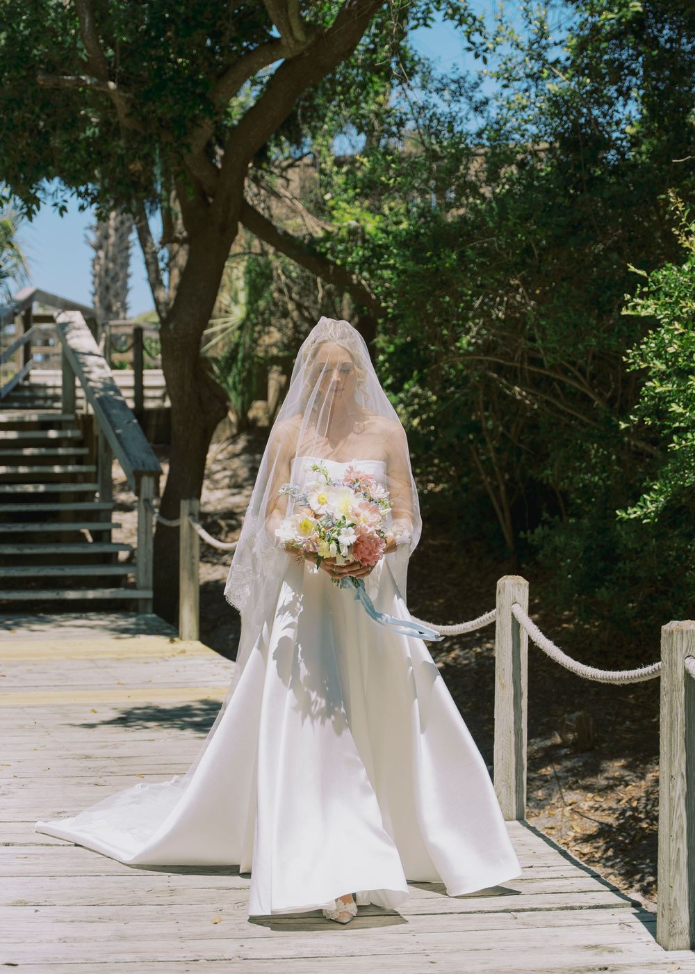 Bride in white wedding gown and veil walks on wooden boardwalk holding floral bouquet outdoors.