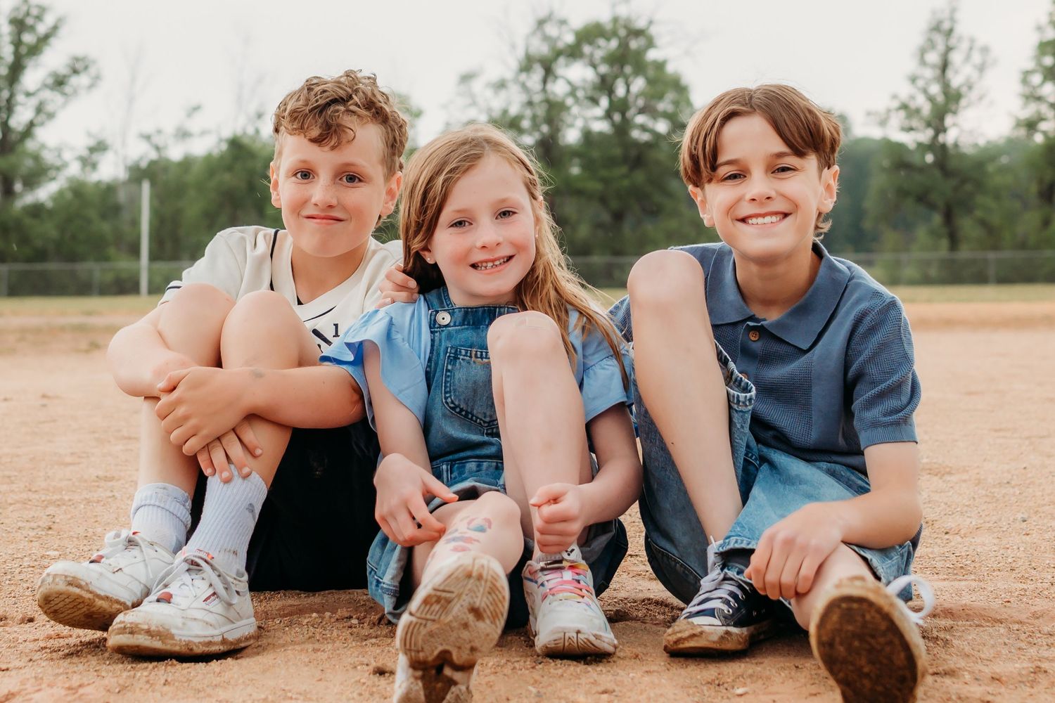 Children sitting in a dusty baseball field wearing casual clothes and athletic shoes.