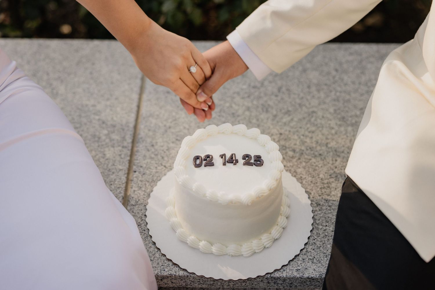 Couples in white outfits cut into a small white wedding cake decorated with numbers and pearl trim details.