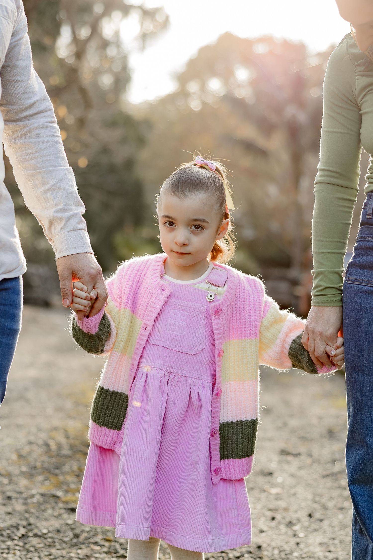 Two adults holding hands with a small child in a pink dress walking outdoors during golden hour.
