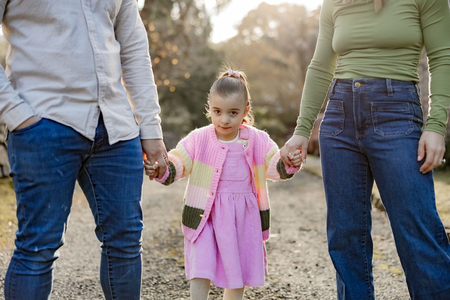 Small child in pink dress holds hands with two adults while walking outdoors during golden hour.