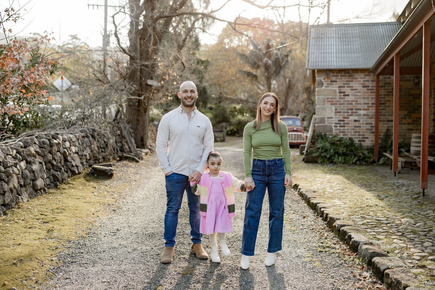 Family stands together on a gravel path outside a rustic brick home with an autumn backdrop and stone wall.