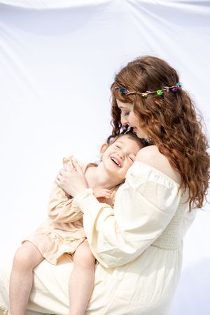 A woman in a cream off-shoulder dress and floral headpiece shares a joyful moment with a laughing child in matching tones.