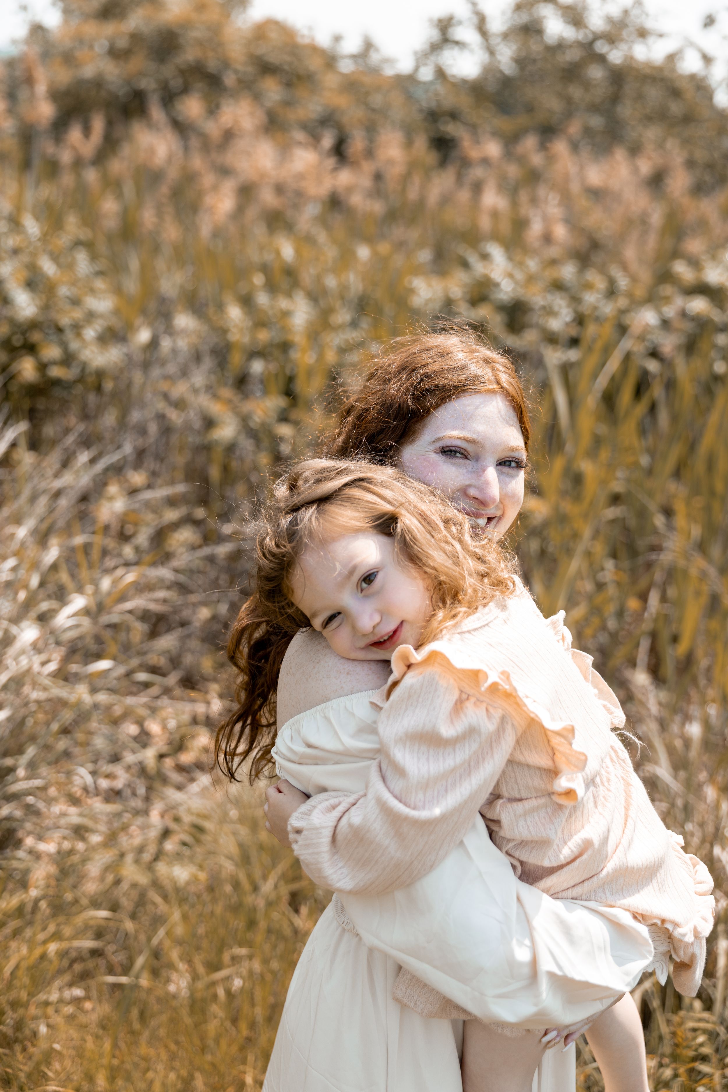 Two figures embrace in a sunlit field wearing white dresses, their red hair blowing gently in the breeze.