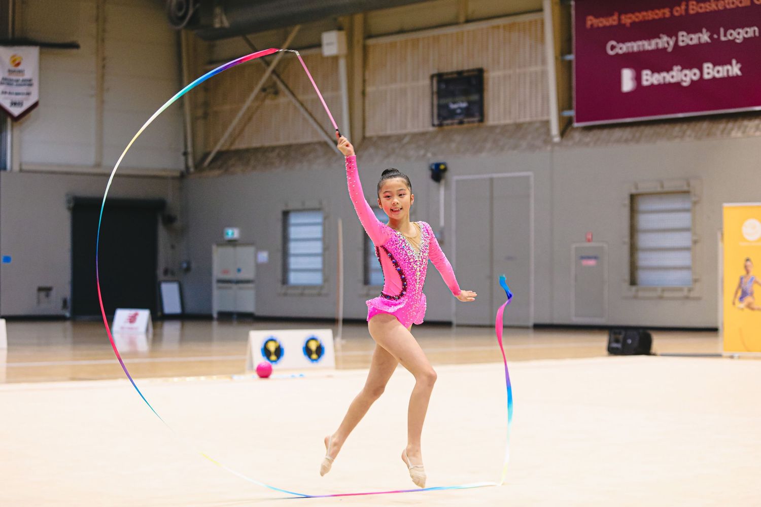 Rhythmic gymnast performs with a pink ribbon and colorful arc during a competition in a gymnasium.