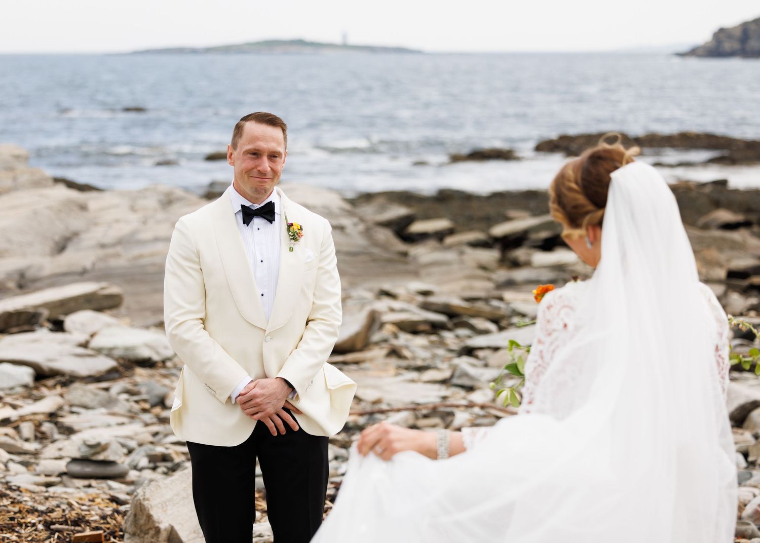 Romantic coastal wedding ceremony on a rocky beach with elegant white attire and ocean backdrop.