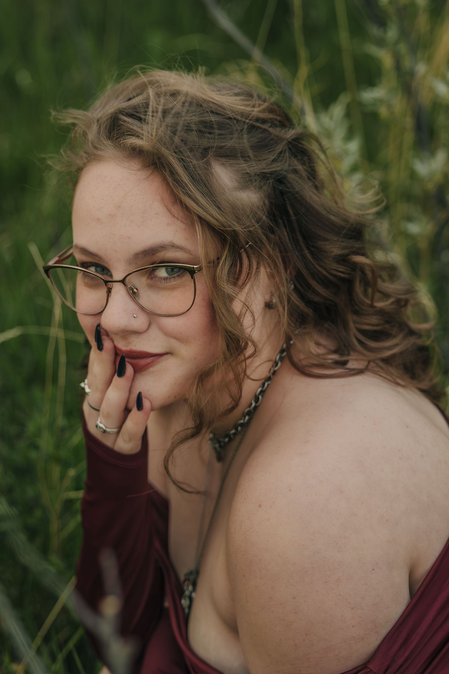 Person in glasses with curly hair wearing burgundy clothing poses thoughtfully against green foliage background, in Linden, Alberta.