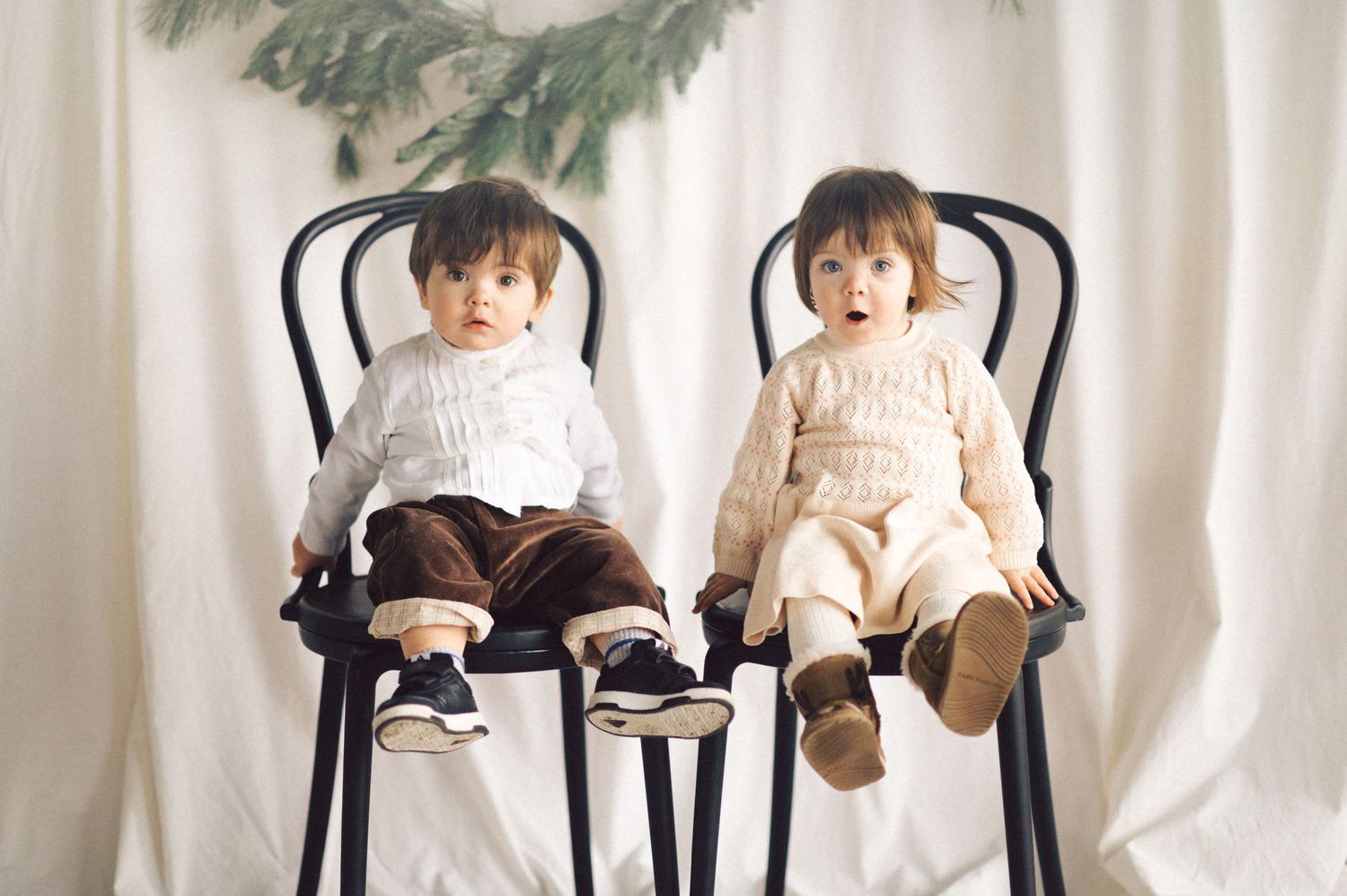 Two babies in matching outfits sit side by side on black bistro chairs against a white backdrop with greenery above.