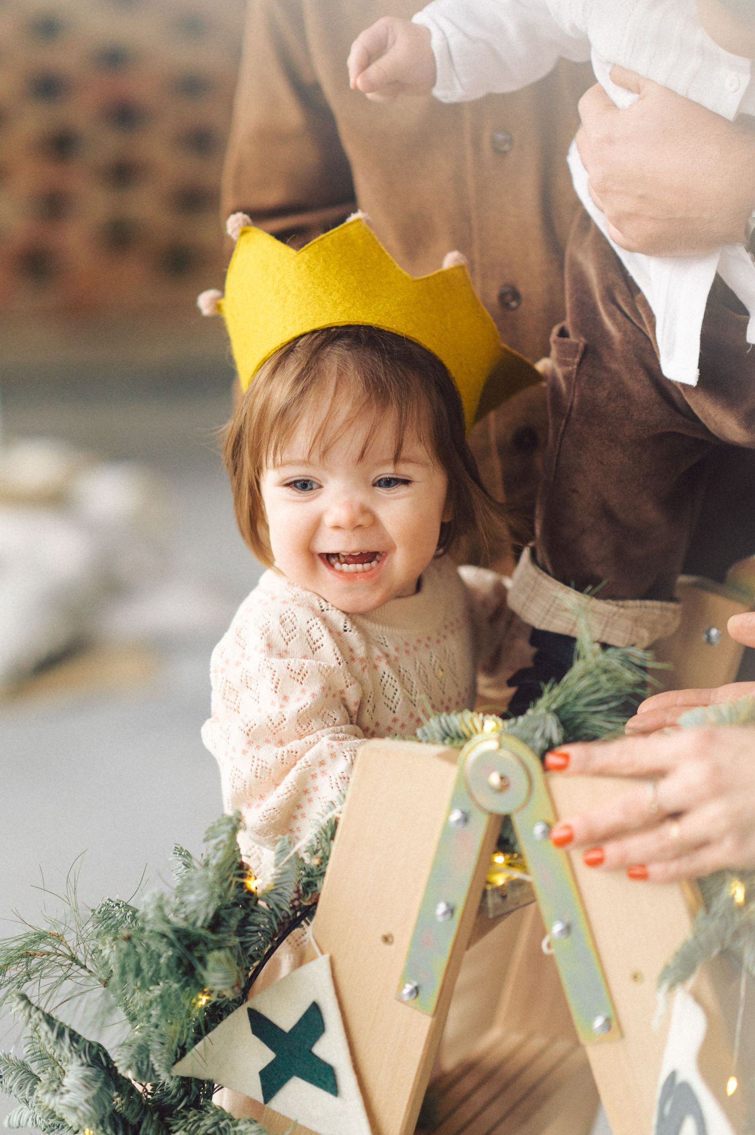A smiling toddler wearing a bright yellow paper crown sits near holiday decorations with greenery.