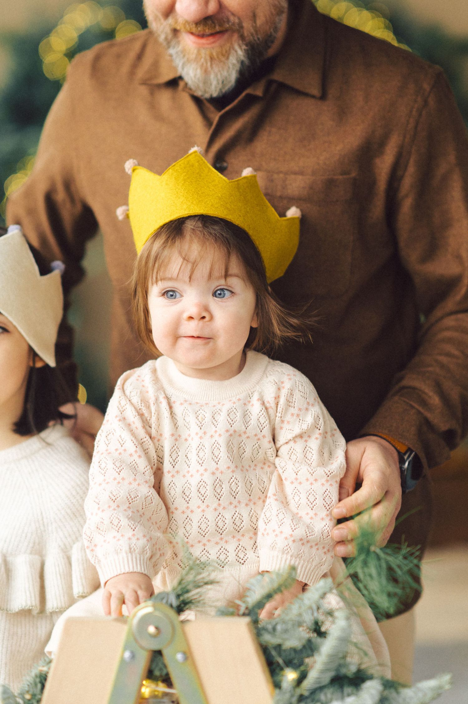 Small child wearing a yellow paper crown and cream sweater sits among holiday decorations.